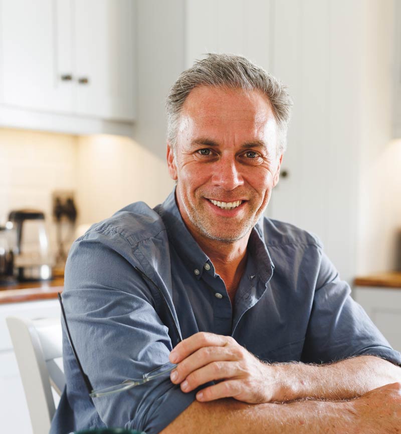 Señor maduro sonriendo sentado, con una cocina al fondo